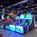 A brightly lit arcade filled with colorful gaming machines sits under ceiling lights. Neon lights illuminate various game stations, creating a playful and vibrant environment. A few red chairs are scattered around the reflective, tiled floor, and a person is observing one of the machines. The atmosphere is lively and cheerful, featuring games with cartoonish designs and engaging themes.